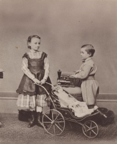 Full-length portrait of Mary Lydia Greene and Howard Greene posing with toys. Mary has a doll in a baby buggy, and Howard has a locomotive placed on a chair. Mary is wearing a plaid under dress with a dark over dress, a necklace, stockings, high-button shoes and a headband in her hair. Howard is wearing knickers (knickerbockers), matching coat, necktie, and boots.