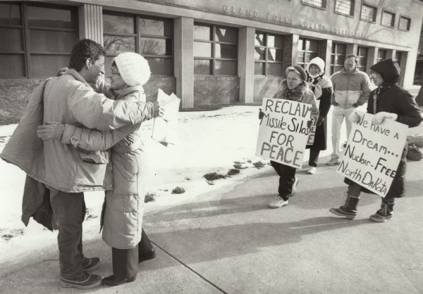 Two people hug on the left while four other people picket on the sidewalk on the right. Two women carry signs, with one sign reading: "Reclaim Missile Silos for Peace," and the other sign: "We have a Dream . . .  Nuclear-Free North Dakota." They are picketing on the sidewalk outdoors in front of the Grand Forks County Courthouse, and are dressed for winter in coats, hats and boots.