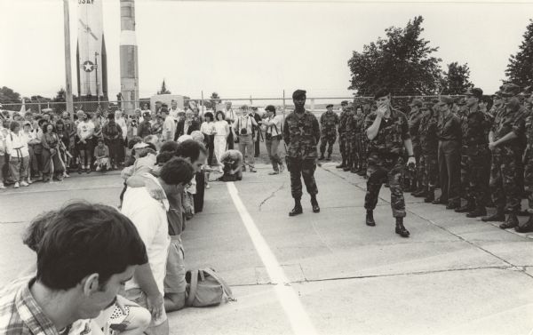 Stratcom action photo, with several protesters kneeling behind a line with soldiers on the other side. In the background are a fence, missile, rocket and trees.