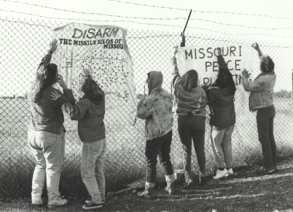 Missouri Missile School photograph of six women hanging banners on a chain link fence topped with barbed wire. One banner reads: "Disarm The Missile Silos of Missouri," and the other reads: "Missouri, Peace, [?]ing"