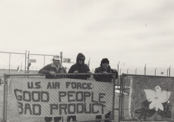 Three people behind a fenced gate with two signs. One reads: "U.S. Air Force: Good People, Bad Product," and the other sign, on the right, has a peace dove over an olive branch. Behind the group are fences topped with barbed wire.