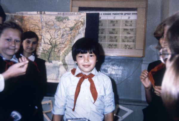 A boy in a classroom wears a red necktie with his school uniform. Other children stand around him also wearing red neckties.