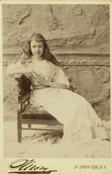 Nina Dousman, age 13, relaxes in a chair for a full-length, cabinet card sized, studio portrait. She has her fingers interlaced and is wearing a light-colored dress with an embroidered waistband. The backdrop is a decorative plaster wall. A fur rug is on the floor.