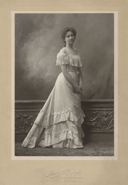 Full-length formal portrait of Virginia Dousman. She is wearing a light-colored, off-the-shoulder dress, with straps. Her hands are clasped. In the background is a chair rail with sculptured panel molding, and above is a painted backdrop. A carpet is on the floor.