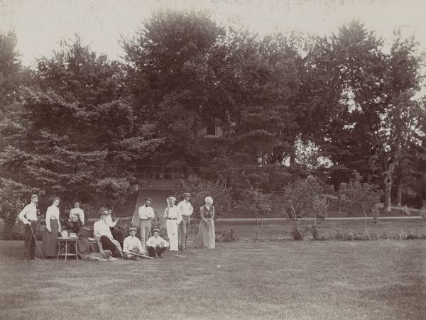 Dousman family entertaining house guests on the lawn at Villa Louis. A table and chairs are set up on the grass and several of the group are holding baseball, tennis and golf equipment. Two dogs sit with the group of people. In the background is the house under trees.