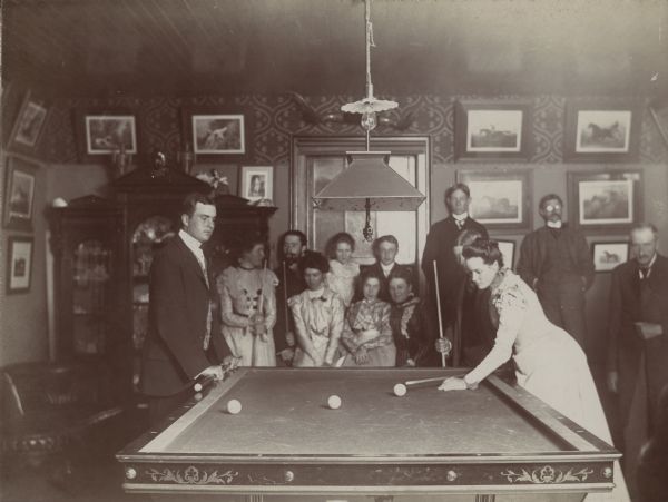 Dousman family and house party guests playing billiards at Villa Louis. A lamp hangs over the table and prints are displayed on the walls. A display cabinet and leather sofa are on the left.