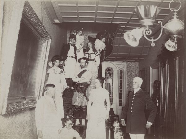 Dousman family and house party guests dressed for a fancy ball. They are posed on the staircase and in the hall and are wearing fancy dress or costumes. One of the people towards the bottom steps is dressed in blackface. A large painting hangs on the left wall.
