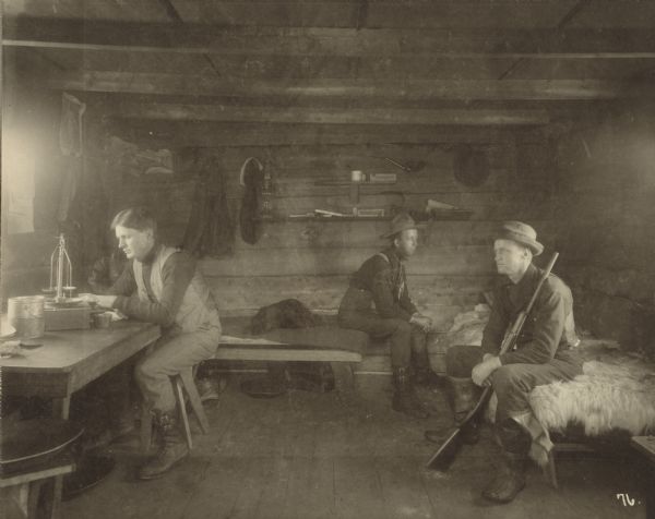 Dr. A.N. Kittelsen (left) seated at the table, his brother Bernhardt Kittelsen (right) seated on a fur covered bed, both of Stoughton, Wis. and Norman Torrison of Manitwoc, Wis. (center) seated on the far bed, assaying gold. Bernhardt is holding a gun. The interior of the building is bare boards. Various items are hung on the wall or resting on shelves.