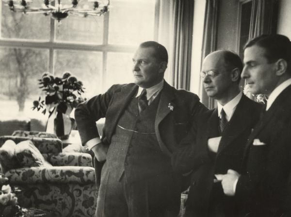 Herman Goering, Louis Lochner and an unidentified man standing in a row indoors in a room with a large window in the background. In the room is stuffed furniture, a chandelier, drapes and a vase of flowers.
