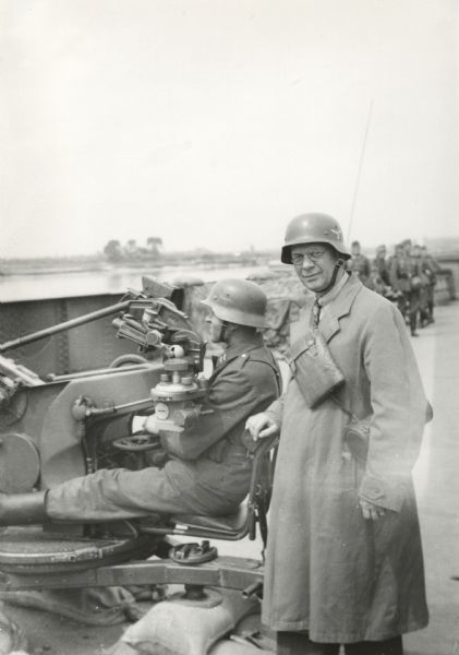 Louis P. Lochner, somewhere in Germany, stands next to a Nazi soldier seated in a piece of artillery. In the background are more soldiers. Lochner is wearing a helmet, long coat and two bags slung around his chest.