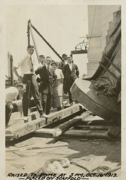 A group of men and women look on as the statue, "Wisconsin," tipped sideways, is lifted from the scaffold. Only the bottom section of the statue is visible. 
