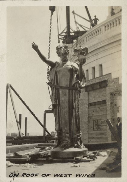 Several men are looking at the statue "Wisconsin" as she rests on a low scaffold on the roof of the West Wing. The statue is wrapped with rope and attached to a chain hanging above from a crane. Text on photograph reads: "On Roof of West Wing."