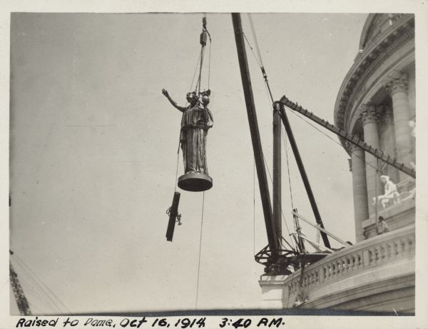 The statue "Wisconsin" in midair being raised from the scaffold on the roof of the West Wing up to the dome. Men are standing on the balcony on the right.