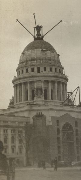 View from street of scaffolding and cranes on the Wisconsin State Capitol used to construct the building. Pedestrians and a carriage are in the foreground.