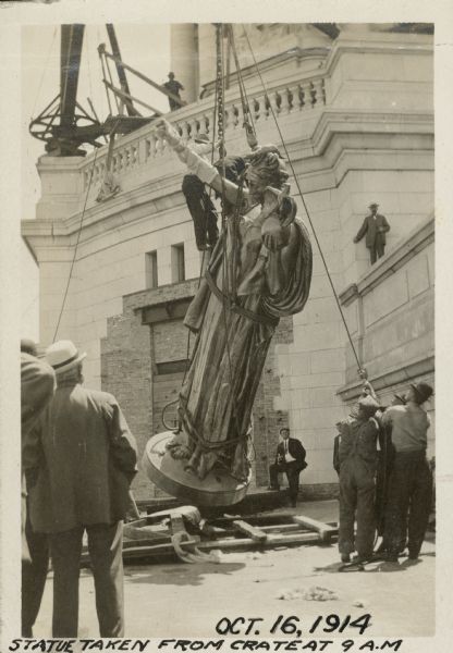 A group of men look on as the statue "Wisconsin" is lifted from the crate using ropes and pulleys. Ropes are wrapped around the statue and the statue's right arm is wrapped to prevent damage. One man is suspended near the chain above the sculpture's.