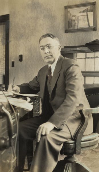 Indoor portrait of Chester P. Gross, seated in a chair at a desk. He is writing in a notebook with a pencil. Gross photographed the Wisconsin State Capitol construction, and the installation of the statue "Wisconsin" as they lifted it to the dome.
