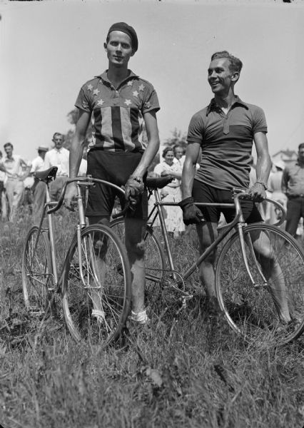 Two bike race competitors pose with their bicycles in a field as an admiring crowd stands behind them. One man wears a "stars and stripes" shirt and a beret.