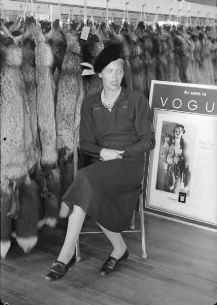 A woman is seated in front of silver fox pelts hanging from pegs at the Fromm Brothers Fur and Ginseng Farm sale. On the right is a poster of an advertisement from <i>Vogue</i> magazine. The woman is wearing a dress, shoes, multiple strands of pearls and a hat.