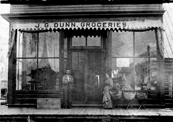 Photographic postcard of the James G. Dunn Grocery, with William John Dunn standing on the left and Maggie (O'Hare) Dunn on the right.<p>Handwritten on the back is: "Corner of Pelham & Randall (Mercer) Rhinelander — Store and apts. — 4-5 Rm. & 2-3 Rm. (over store.) James Geo. Dunn Grocery - NOTE: His middle name was Gerald, not George. Wm. John (son) standing outside. (James Vs father). Woman may be J.G's wife Margaret O'Hara (O'Hare) Dunn or one of his daughters. Probably very late 1800s or early 1900s — Grandma (Margaret) had a millinery shop in left front corner (of the store)." The building was torn down in October of 1975 to make way for the new Bergman-Kopp Chiropractic Centre.