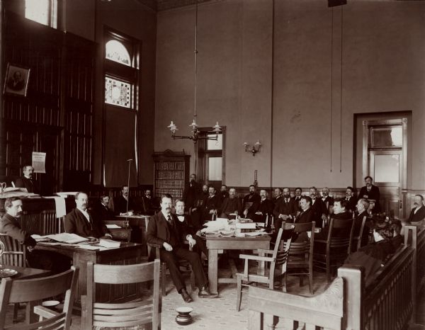 Wisconsin Supreme Court in session at the second hearing of the trial of Emery, Lord and Houston for murder. The case also had two Circuit Court hearings.<p>Identifications: On the bench, Judge Charles V. Bardeen, later (1898-1903) Chief Justice; foreground, left to right: Unknown man; William H. Mylrae, later (1895-99) Attorney General; B.A. Goggins, District Attorney (who wears an artificial left leg, due to a boyhood logging accident); George R. Gardner, assistant to the District Attorney (who died during this trial); on the right side of the table: George Williams, defense attorney; Neal Brown, defense attorney, later a State Senator from Wausau and a prominent orator; Lord, one of the defendants, later hung for the murder. A Bailiff stands by each of the doors. The names of the jury would be obtainable from the record. The man directly behind the upright rod, to the right of the bench, may be one of the other defendants. These identifications have been provided by T.W. Brazeau of Wisconsin Rapids, Wis., a partner of B.A, Goggins.</p>