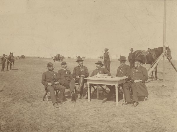 Outdoor group portrait of six men sitting around a table in military uniform in an open field. On the table are pens, an inkwell, two cigar boxes and papers. In the background are scattered groups of men, horses and horse-drawn vehicles. On the right is the bottom of a flagpole. On the horizon are tents and a flag. Names, (l to r), C.E. Burmester, S.B. Jones, A. Allee, J.S. Clarkson, Commander H.C. Russell, and J.D. Miles.