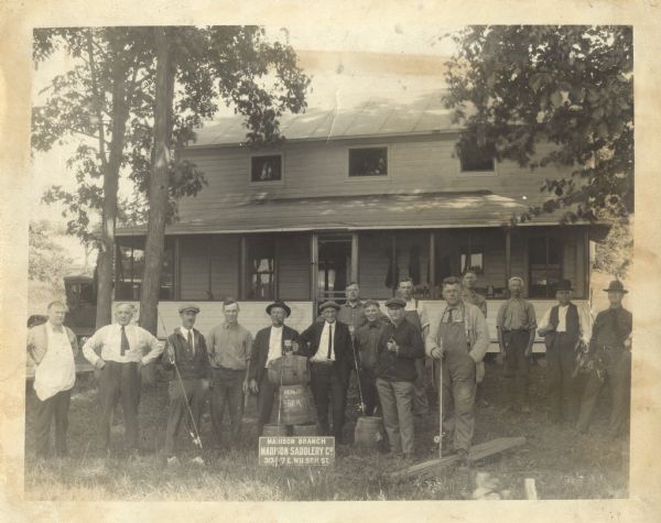 Outdoor group portrait of the Madison Branch of the Madison Saddlery Company located at 313-317 E. Wilson Street. The event may be a recreational gathering for the employees, with beer and fishing. In the background is a house with a large covered porch and trees. Fifteen men pose standing with five fishing poles, two men are smoking pipes, and one is smoking a cigarette. There are four kegs of beer stacked in the center. One keg has "REAL BEER" printed on it. The man in the center holds a stein of beer on the top keg. An automobile is parked alongside the building on the left.