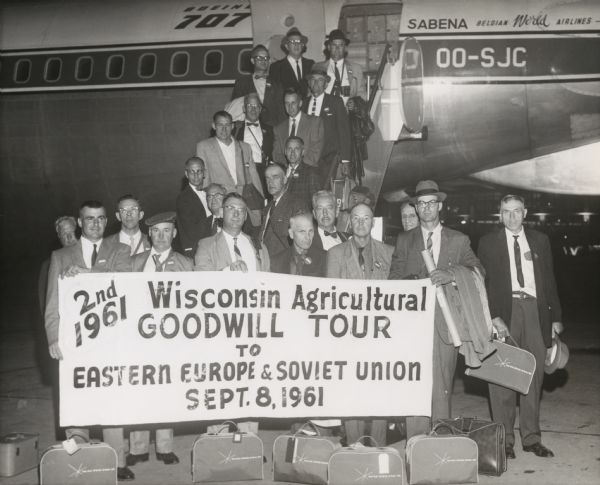 Outdoor group portrait of 22 men with their matching carry-on bags. The men are posed on the gangway and the tarmac in front of an airliner. The airplane is a Boeing 707 of the Belgian National Airline, SABENA Belgian World Airlines. They are holding a banner that reads: "2nd 1961 Wisconsin Agricultural Goodwill Tour to Eastern Europe & Soviet Union Sept. 8, 1961."