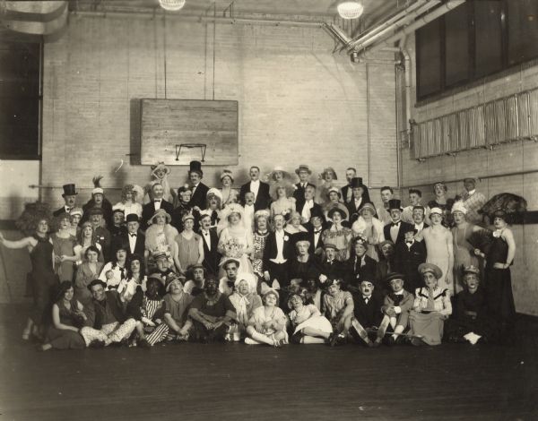 A womenless group wedding portrait taken in a gymnasium. The large group of men are all dressed theatrically in male and female apparel. Some of the men are in blackface. The bride and groom are in the center.