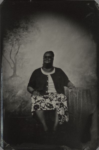 Modern tintype, full length, formal portrait, of Angela R. Davis, Development Officer, Wisconsin Historical Foundation. She is seated in a chair with her arm resting on a table, in the background is a painted backdrop.