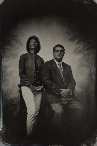 Modern tintype, full length, formal portrait, of Scott and Kate Neitzel. Scott is the Secretary of the Wisconsin Department of Administration for the State of Wisconsin. Kate is on the Wisconsin Chamber Orchestra Board of Directors. He is seated in a chair with his arm resting on a table and she is standing in front of a small table. In the background is a painted backdrop.