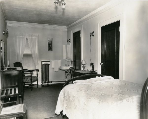 A room at the Eugene Hotel, with a bed, dresser, tables, luggage rack, chairs, lamps and a telephone. There is a sink in the corner of the room. The hotel was heated with hot water registers.<p>The hotel was located on the southwest corner of the square.</p>