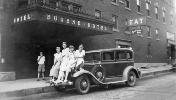 Four young women wearing dresses are posing while sitting on the hood of an automobile parked at the curb in front (south side) of the Eugene Hotel. A boy is standing on the sidewalk behind them eating an ice cream cone. The marquee overhead spells out "Hotel" and Eugene-Hotel." Over another door is a sign that reads, "EAT, Eugene Coffee Shop."<p>The hotel was located on the southwest corner of the square.</p>