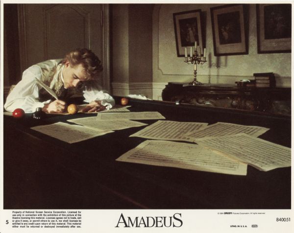 Small color lobby card (number 5) for the film <i>Amadeus</i>. Tom Hulce, as Amadeus, works on a score on a billiard table. There are pages of the score scattered on the table.