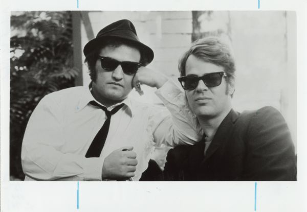 Outdoor portrait of John Belushi and Dan Ackroyd as Jake and Elwood Blues from the film <i>The Blues Brothers</i>. They are both wearing dark sunglasses. Belushi wears a hat and has his elbow resting on Ackroyd's shoulder.
