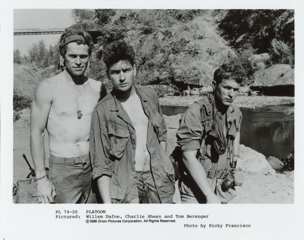 Willem Dafoe, Charlie Sheen and Tom Berenger looking at the camera and standing next to each other in character for the film <i>Platoon</i>. In the background is a river.