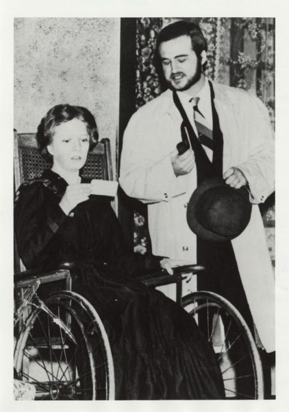 John Belushi performing a scene with Virginia Newcomb from the play <i>Night Must Fall</i> at UW-Whitewater. Newcomb is sitting in a wheelchair and Belushi is standing next to her. He is holding a hat in one hand and a pipe in the other.