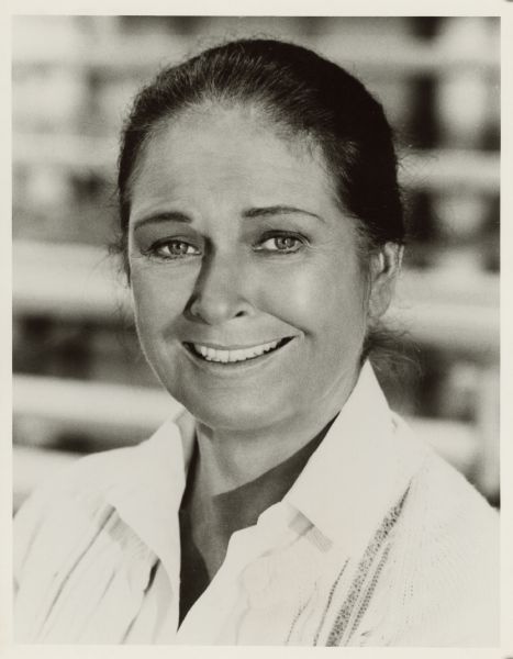 Head and shoulders portrait of actress Colleen Dewhurst. She is smiling and looking at the camera. Her hair is pulled back and she is wearing a white sweater over a white blouse.