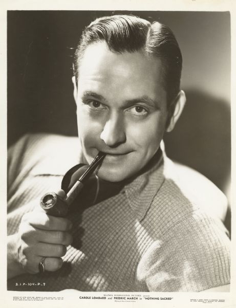 Fredric March, looking directly at the camera, poses with a pipe in his mouth for a publicity photograph for the film <i>Nothing Sacred</i>.