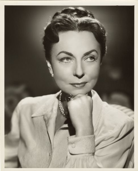 Publicity photograph of actress Agnes Moorehead. She is resting her chin on her hand and is looking off to the side with a slight smile. She is wearing a pearl necklace and earrings, and a large cabochon ring on her left ring finger.