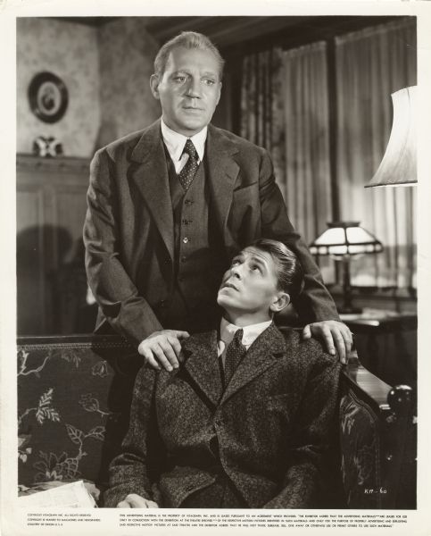Pat O'Brien, as Knute Rockne, standing behind Ronald Reagan who is playing George Gipp, in the film <i>Knute Rockne All American</i>. Reagan is sitting on a sofa and is looking up at O'Brien who has his hands on Reagan's shoulders.