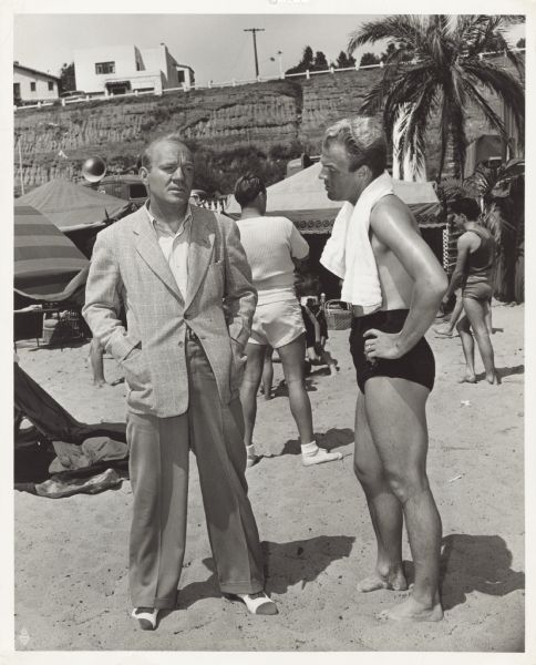 Pat O'Brien talks with Bill Marshall as they stand on the beach on the set of <i>Knute Rockne All American</i>. O'Brien is wearing a suit coat and dress pants and Marshall is wearing a bathing suit with a towel draped over his shoulders.