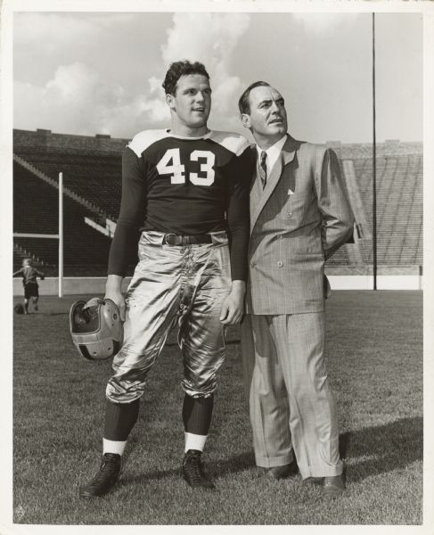 Pat O'Brien stands next to his son Johnny who plays football for Notre Dame University. Pat is wearing a suit and necktie while Johnny is wearing his football uniform.