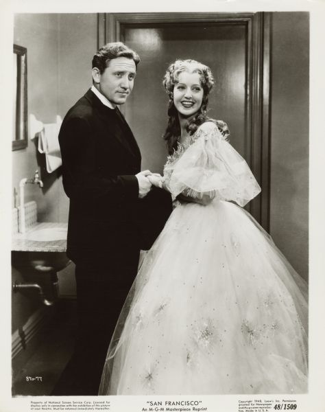 Spencer Tracy and Jeanette MacDonald standing and holding hands in a scene from the film <i>San Francisco</i>. They are looking off to the right. MacDonald wears a full-length off-the-shoulder white dress decorated with stars.