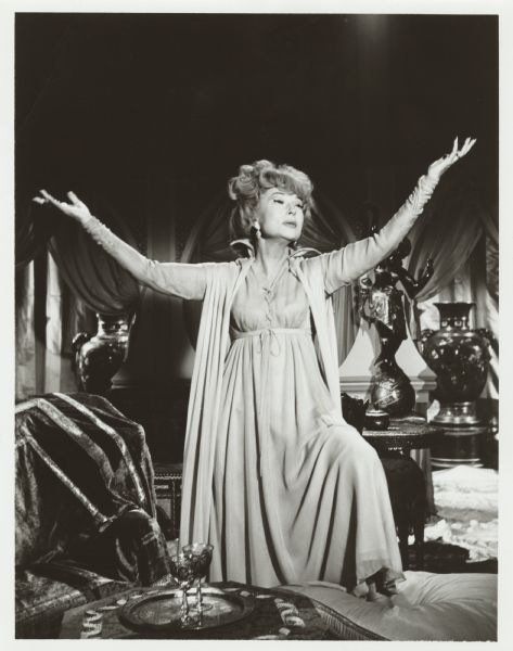 Portrait of Agnes Moorehead as Endora in a scene from the TV show <i>Bewitched</i>. She is standing with one foot on a large cushion and is holding up her arms while looking up towards the ceiling.