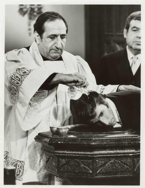 Henry Winkler, as Fonzie, gets baptized by Al Molinaro who plays Father Delvecchio, the twin brother of Al Delvecchio on the TV show <i>Happy Days</i>. Tom Bosley is standing in the background.