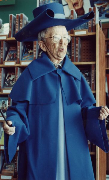 Three-quarter length portrait of an elderly Meinhardt Raabe wearing a costume similar to the one he wore in <i>The Wizard of Oz</i> as the coroner of Munchkin Land. A shelf of videotapes is behind him. 