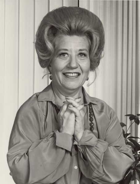 Waist-up portrait of actress Charlotte Rae. She is smiling and standing with her hands folded in front of her.  