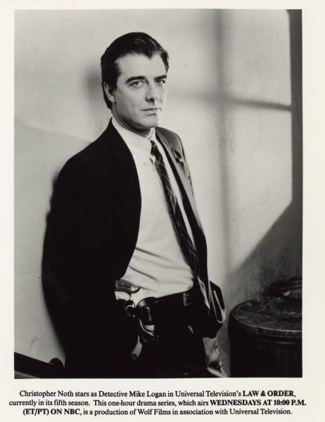 Publicity photograph of Chris Noth for the television series <i>Law & Order</i>. He is standing with his hands in his pants pocket while leaning against a wall. His jacket is open on the right side with his handgun in a holster at his waist.