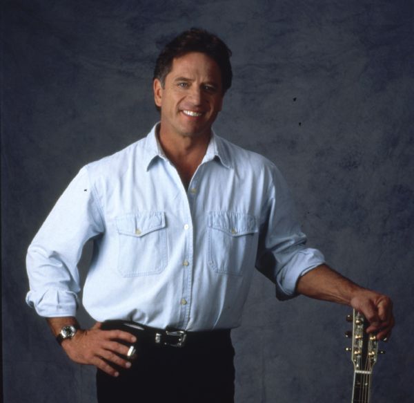 Three-quarter length portrait of Tom Wopat, taken for the television show <i>Prime Time Country</i>. He is standing and has his left hand on the top of a guitar and has a pick on his right hand.