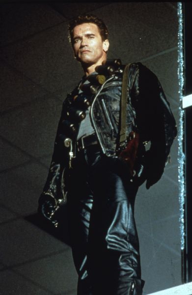 Full-length standing portrait of Arnold Schwarzenegger. He is dressed in black leather and has ammunition and weapons strapped onto his chest and shoulder.